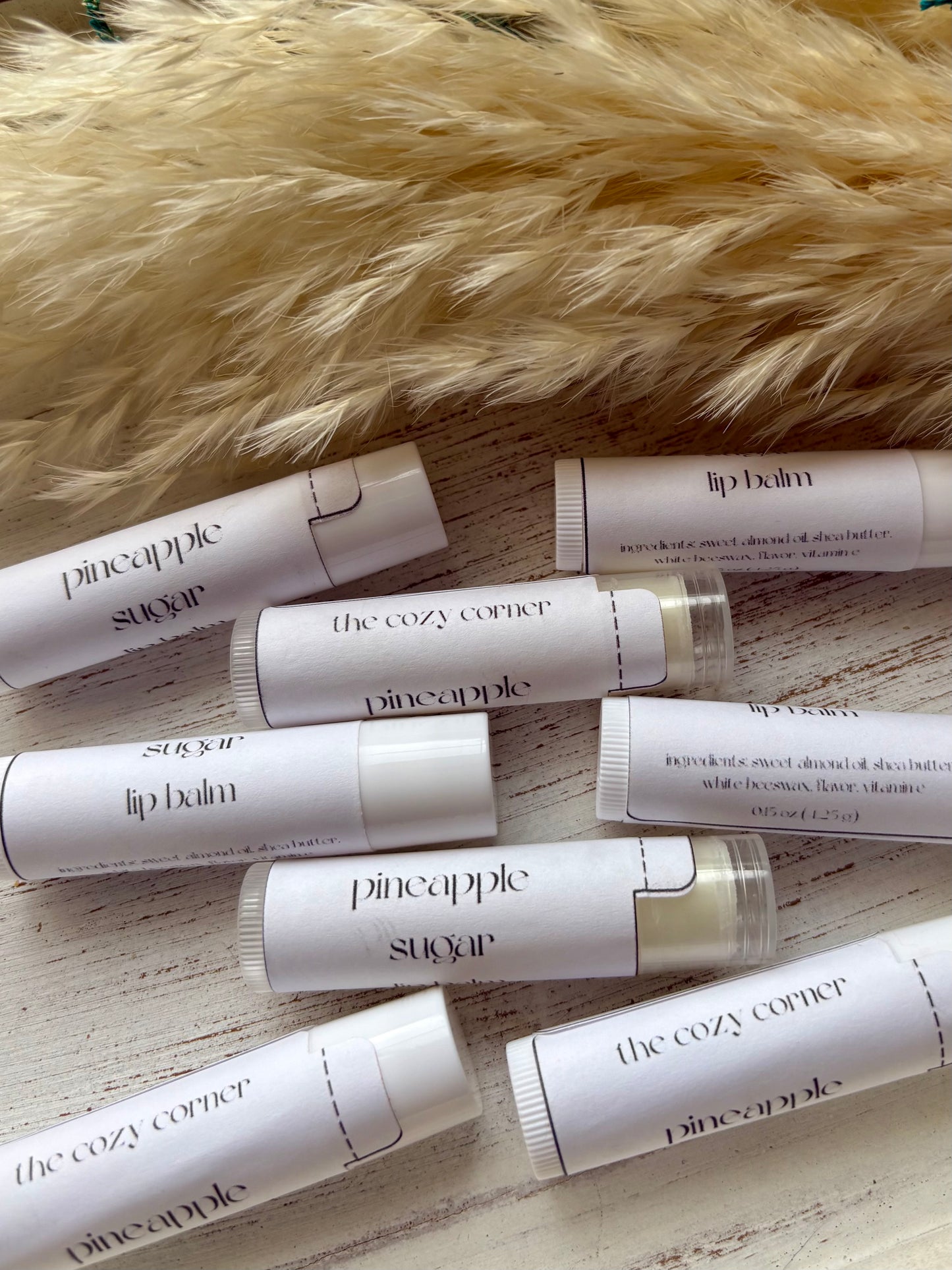 Multiple lip balms from brands like 'pineapple sugar' and 'the cozy corner' on a wooden surface with a fur background.