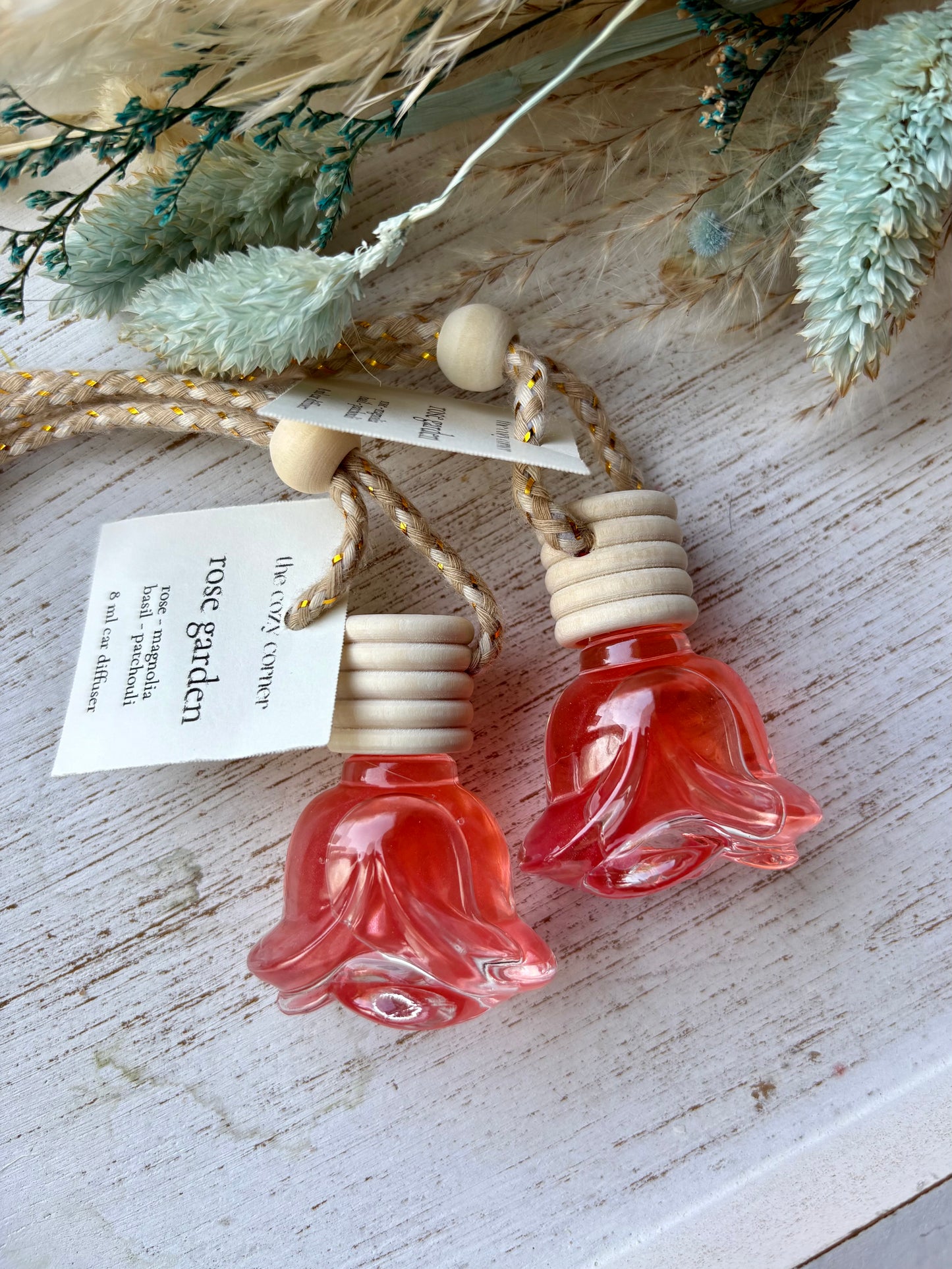 Two small red flower-shaped bottles with wooden caps on a rustic wooden surface with dried plants.