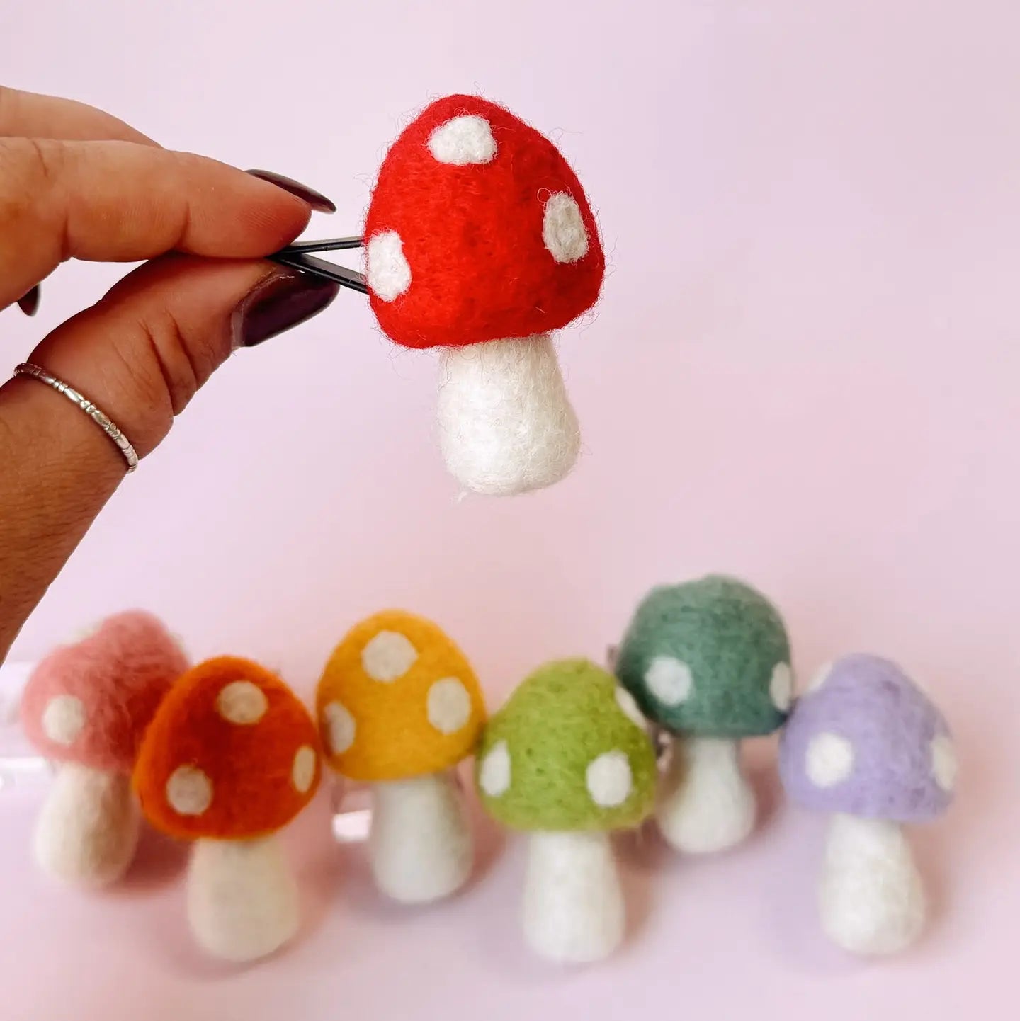 Colorful felt mushrooms with a hand holding one above them on a light pink background
