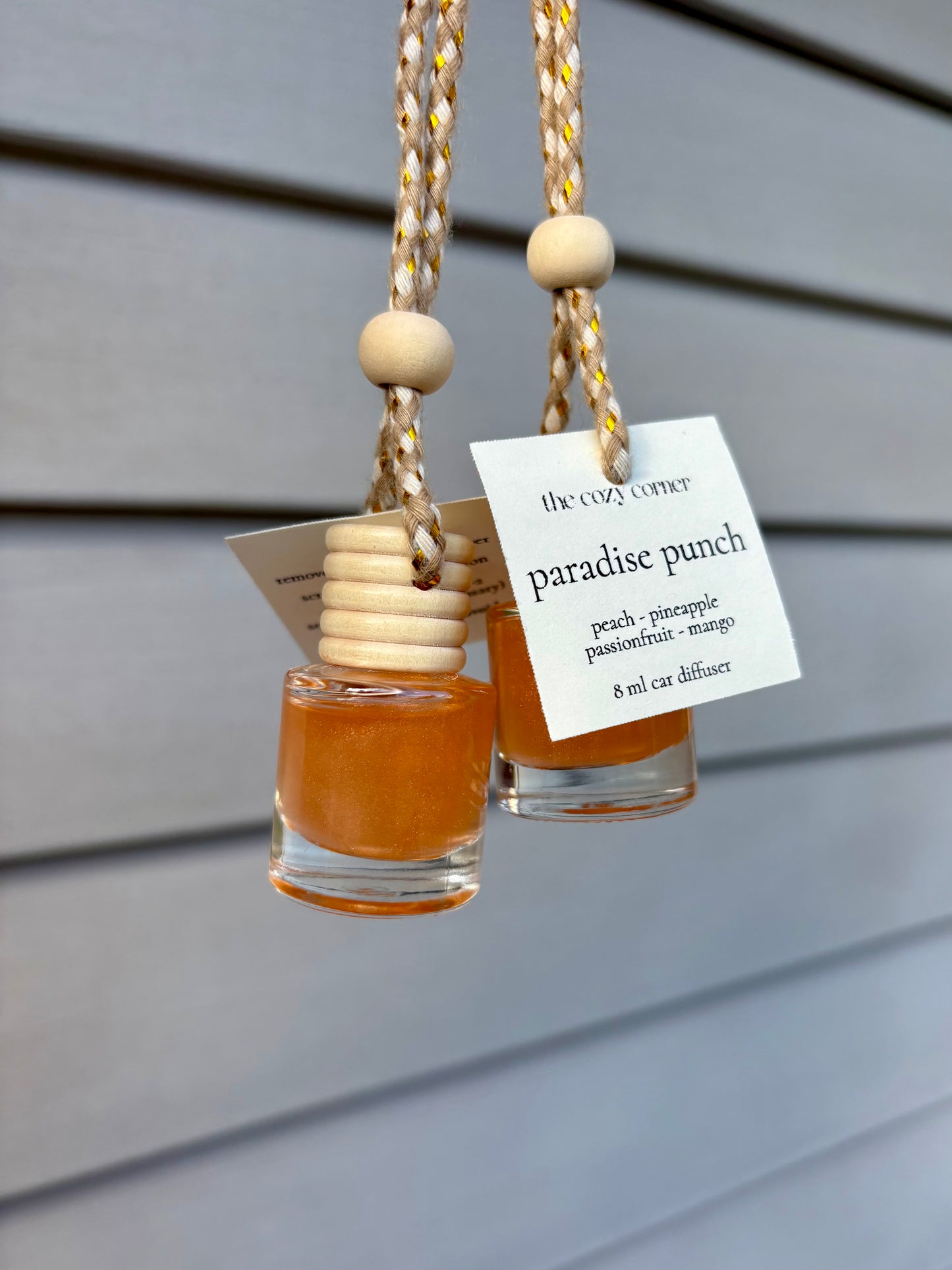 Two small glass bottles with wooden beads hanging against a gray background, labeled 'Paradise Punch'.