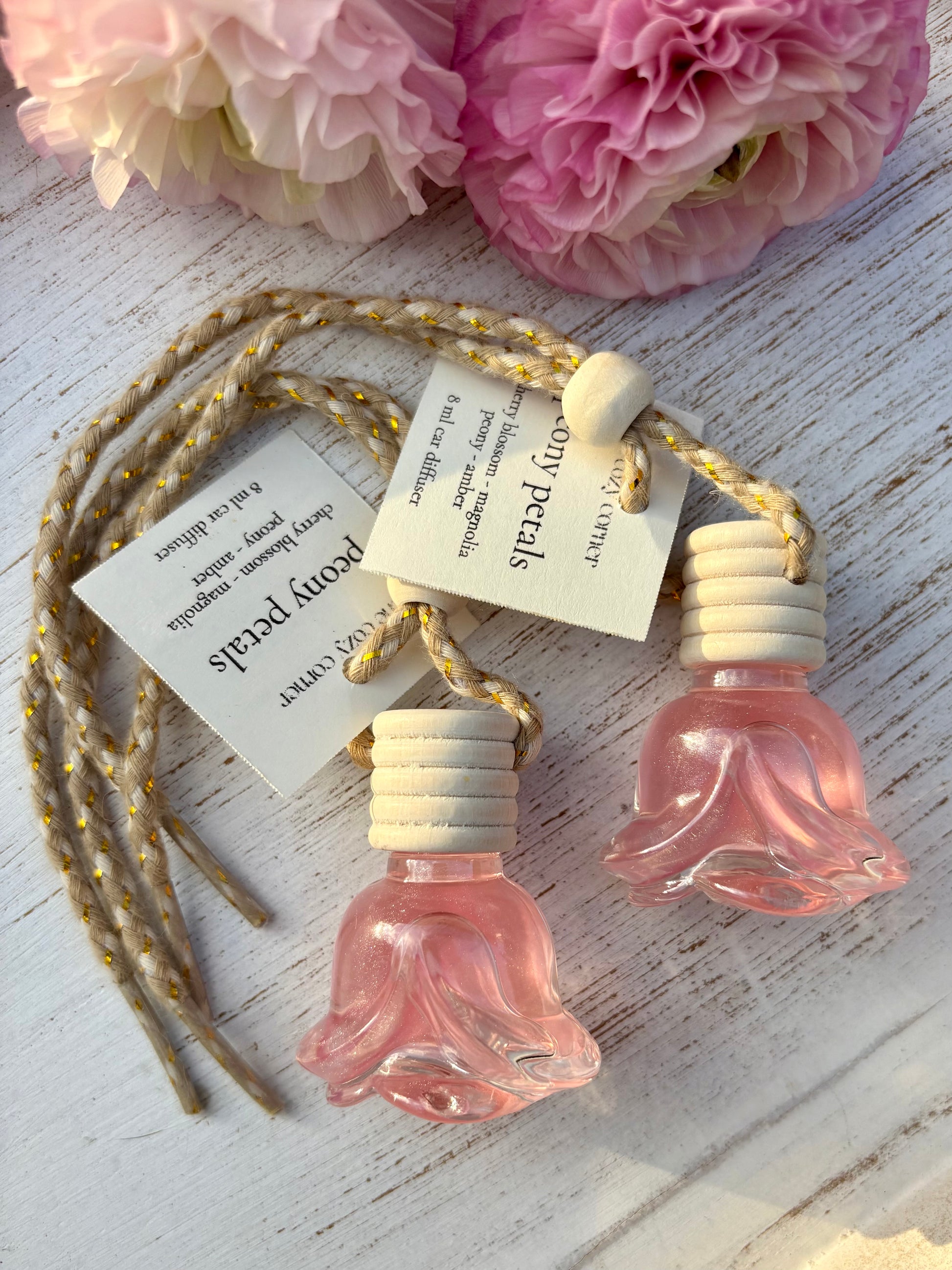 Two pink floral perfume bottles with a tag on a wooden surface with flowers in the background.