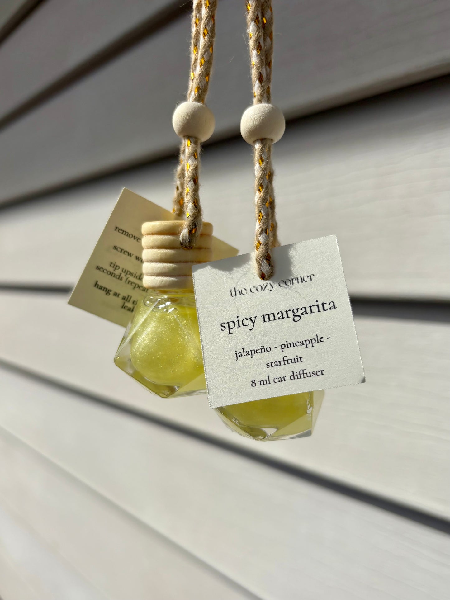 Two small bottles of 'Spicy Margarita' car diffuser with hanging strings on a wooden surface.