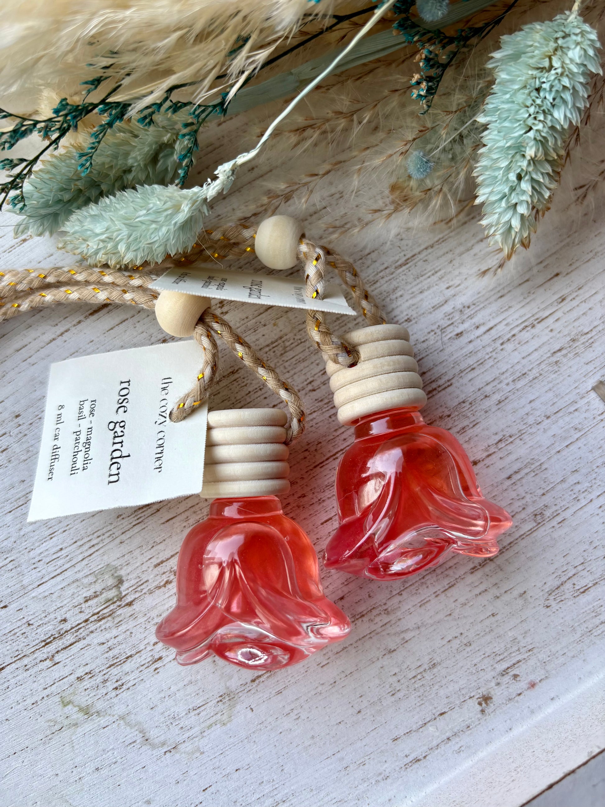 Two small glass bottles shaped like flowers with wooden caps on a rustic surface with dried plants.