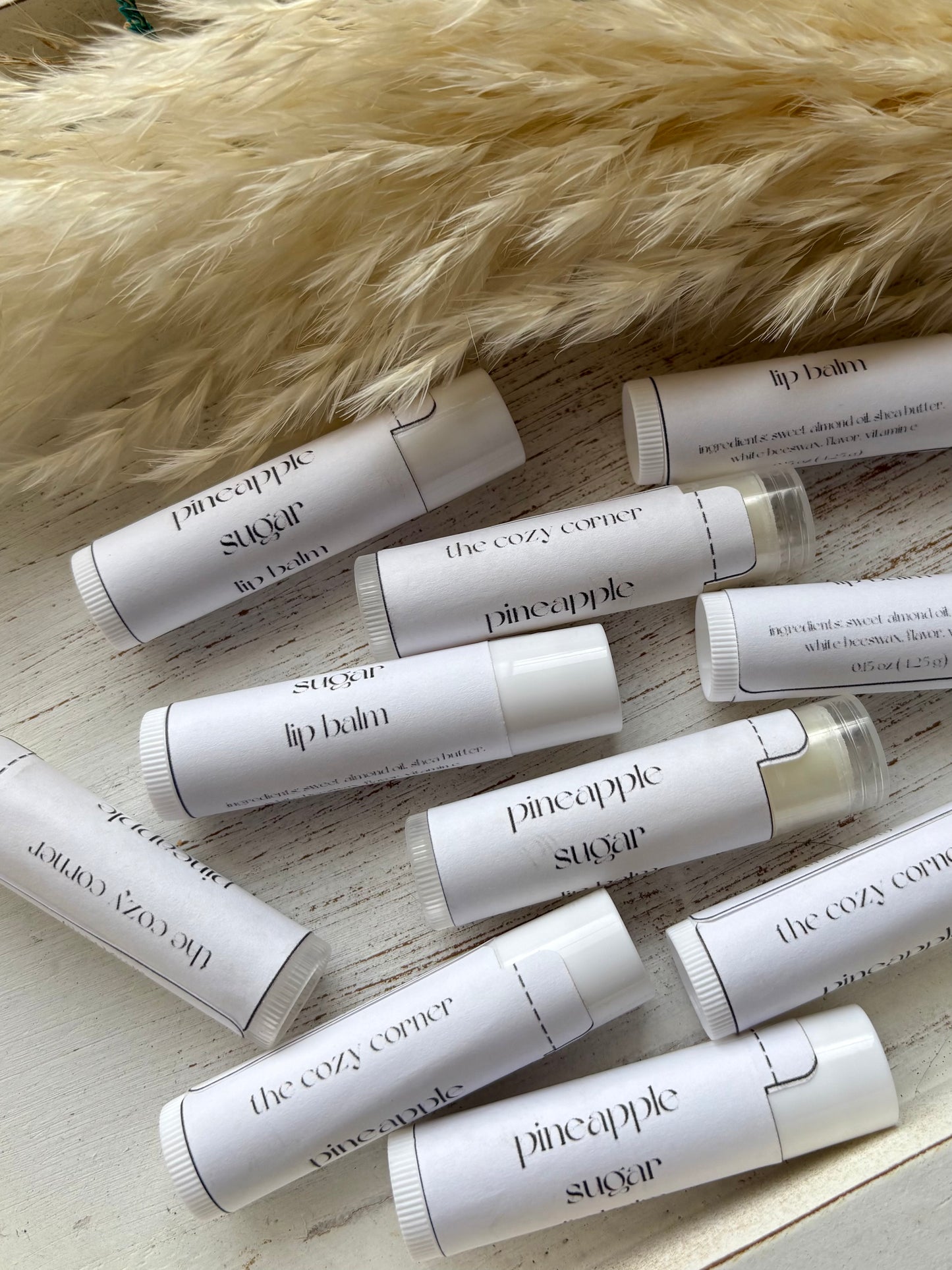 Multiple lip balm tubes from 'The Cozy Corner' and 'Pineapple Suavecito' on a wooden surface with a fur background.