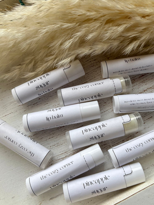 Multiple lip balm tubes from 'The Cozy Corner' and 'Pineapple Suavecito' on a wooden surface with a fur background.