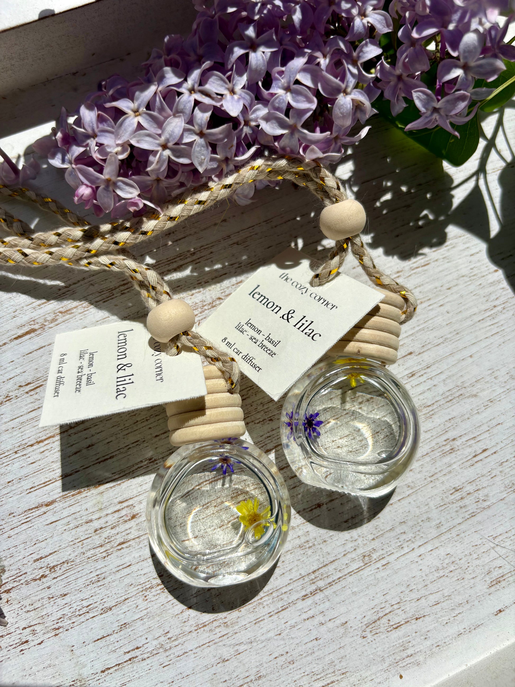 Two glass containers with lemon and lilac scents on a wooden surface with flowers in the background.